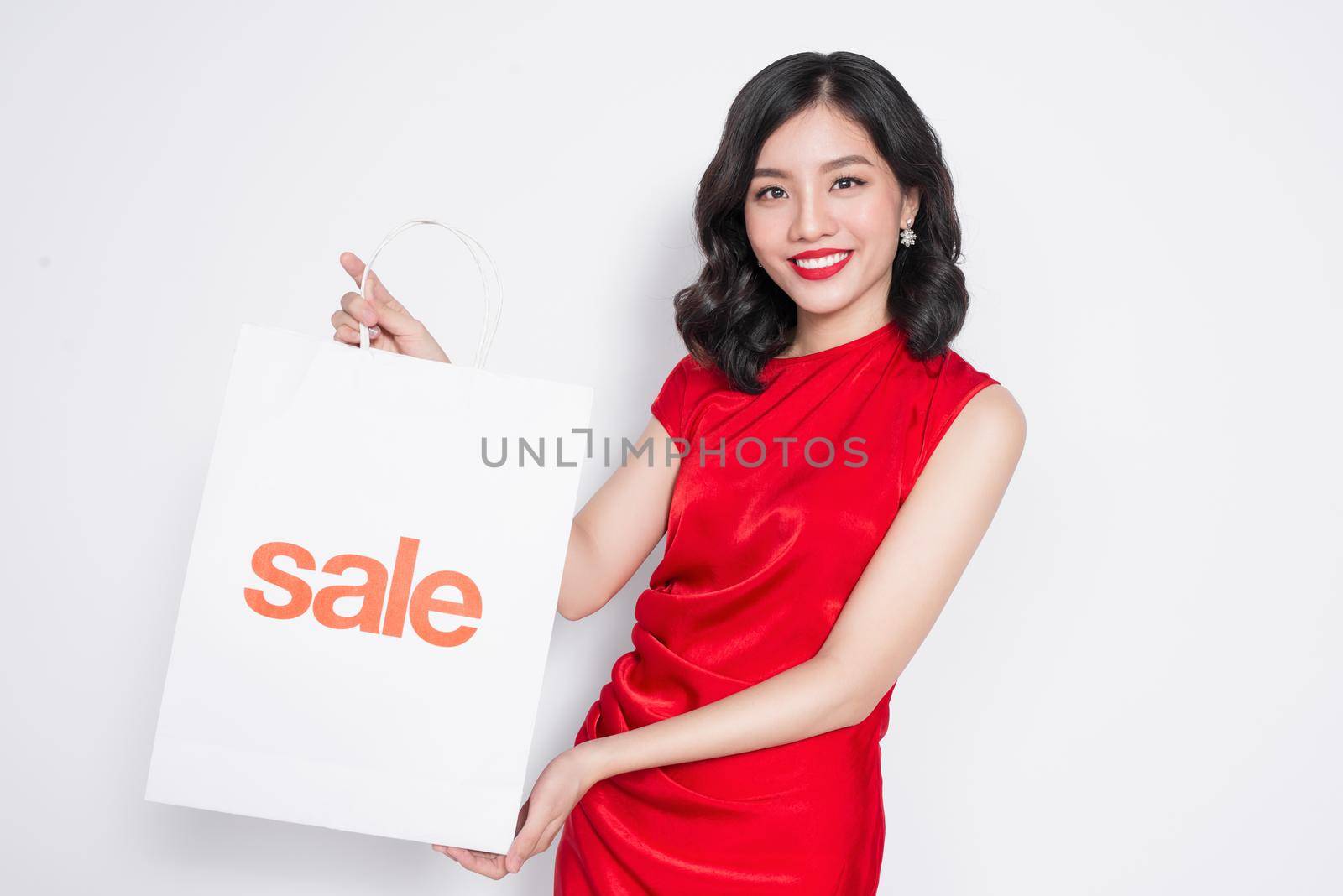 Beautiful asian woman wearing a red dress with shopping bag standing over white.