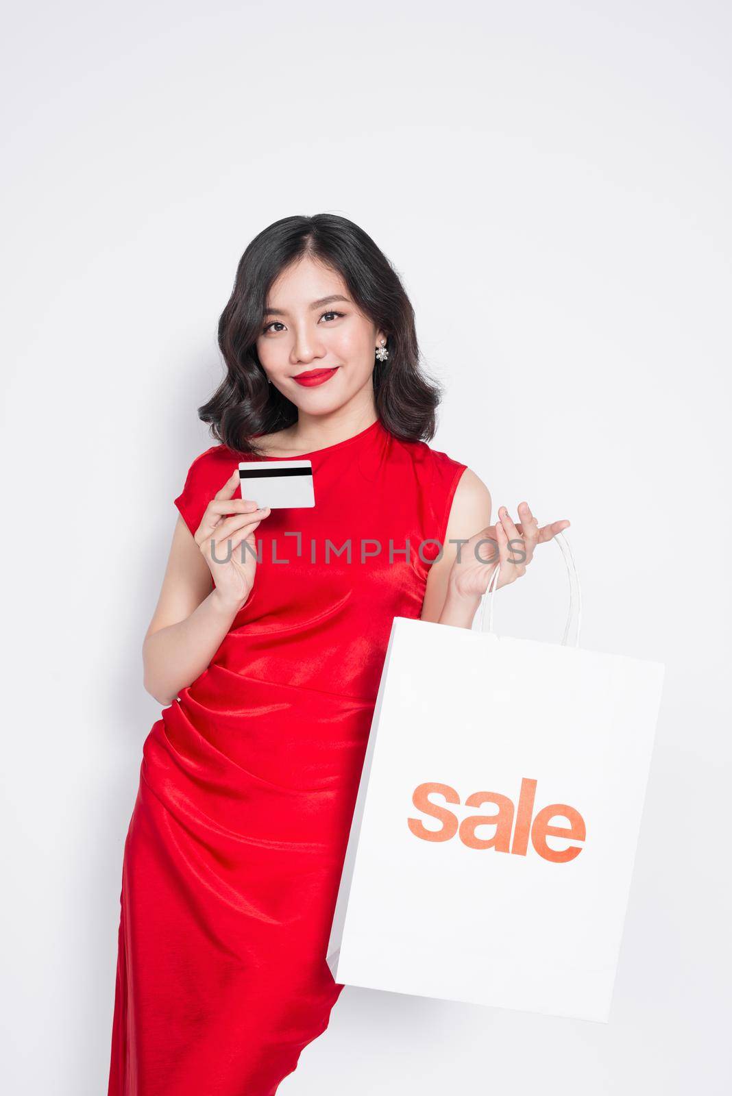 Beautiful asian woman wearing a red dress with shopping bag and holding credit card standing over white.