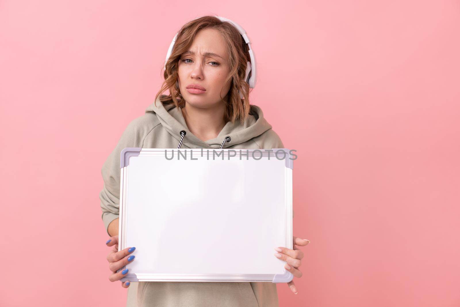 Woman holding empty blank board over pink background Sad unhappy Caucasian female 30 years old dressed headphones oversize hoodie hold blank white board sign Studio shoot Advertising Presentation