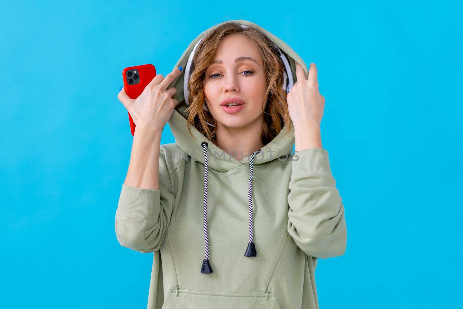 Happy teeth smile woman listen music headphones holding smartphone in hand looking camera Caucasian female enjoy podcast or audio books dressed oversize hoodie blue background close up portrait