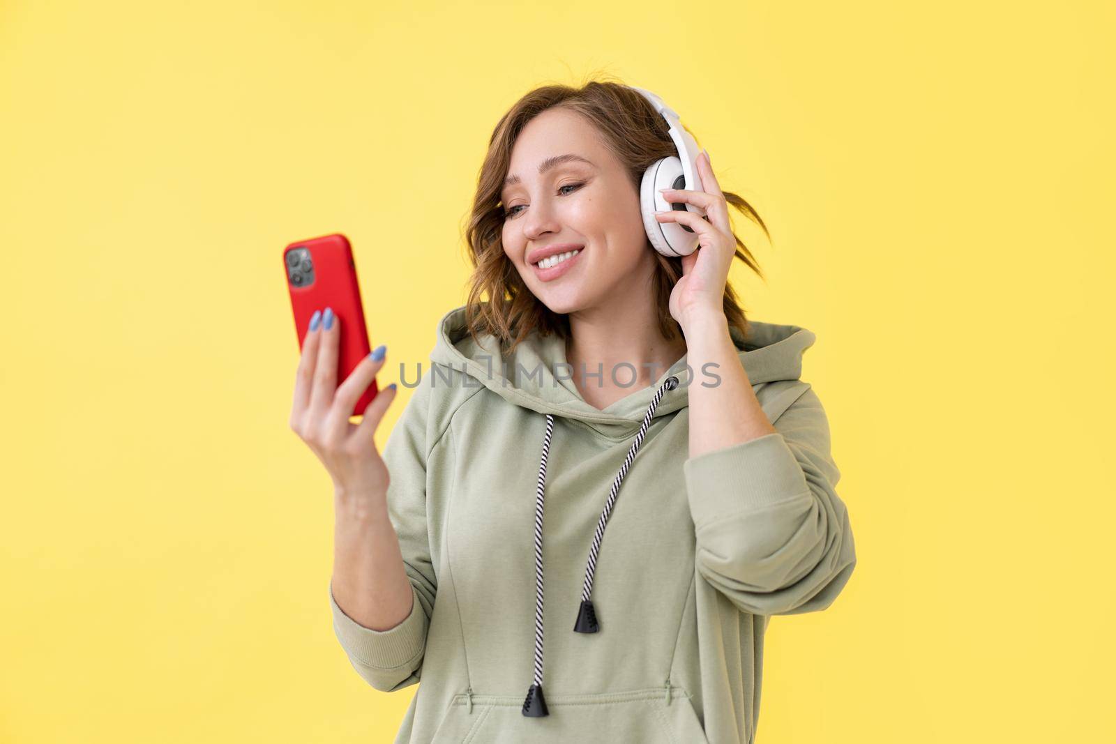 Happy teeth smile woman listen music headphones holding smartphone in hand looking screen Caucasian female enjoy podcast or audio books dressed oversize hoodie yellow background close up portrait