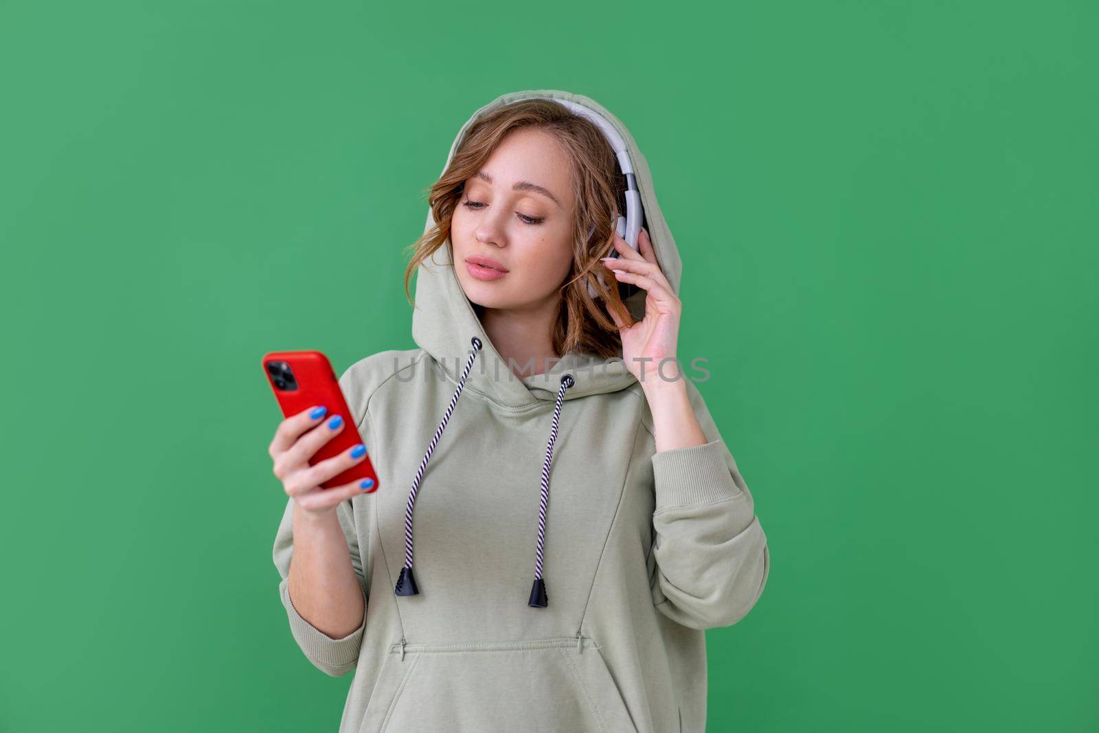 Happy teeth smile woman listen music headphones holding smartphone in hand looking screen Caucasian female enjoy podcast or audio books dressed oversize hoodie green background close up portrait