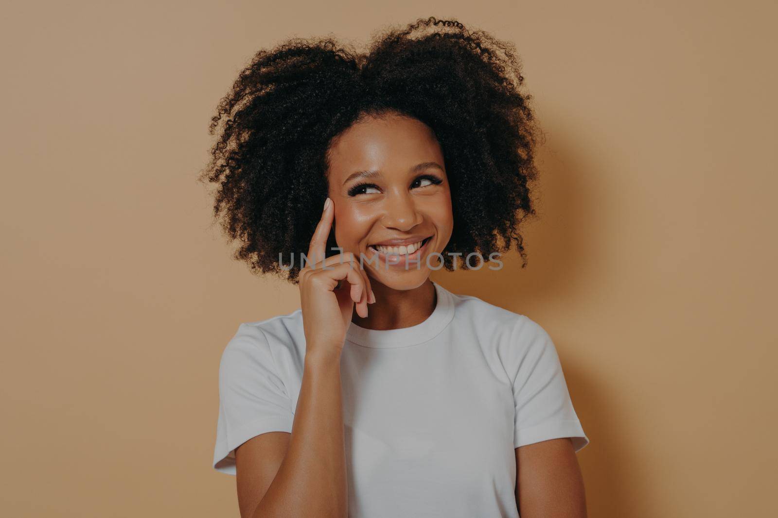 Keep it in mind. Portrait of young positive pensive african woman with curly hair touching temple with forefinger and smiling cheerfully, having good idea, posing isolated over beige background