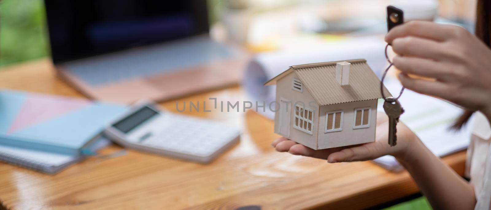 mortgage, real estate and property concept - close up of hands holding house model and home keys with financial working space background