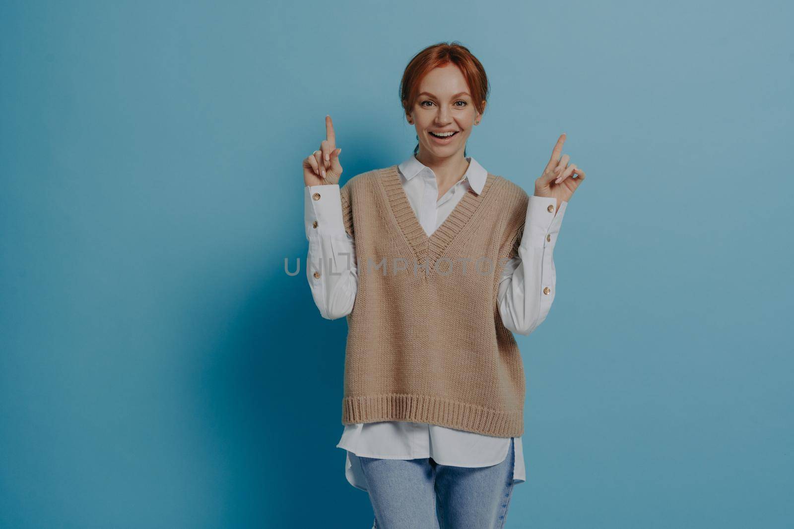 Happy stylish red-haired woman in casual outfit showing up with forefingers at copy space and smiling, isolated on blue, offering something, cheerful ginger female indicating at discount or offer