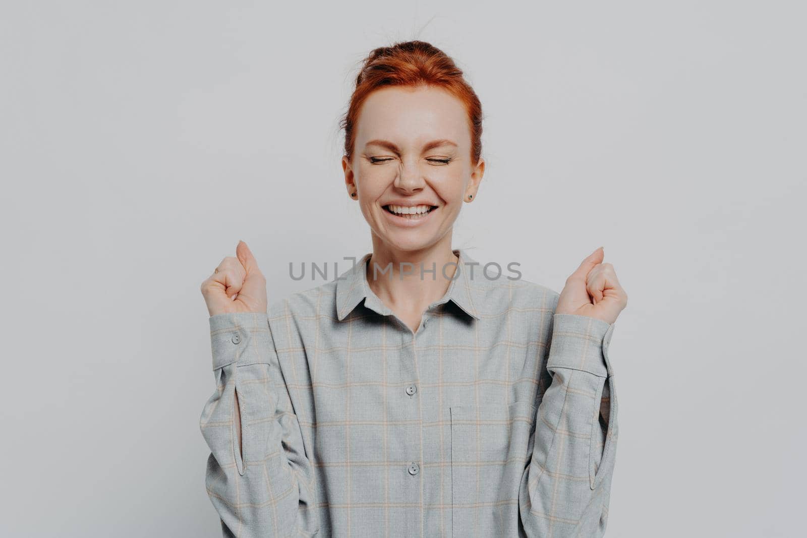 Excited overjoyed young ginger female raising hands with clenched fists and keeping eyes closed, making yes gesture while posing isolated on grey studio background. Success concept