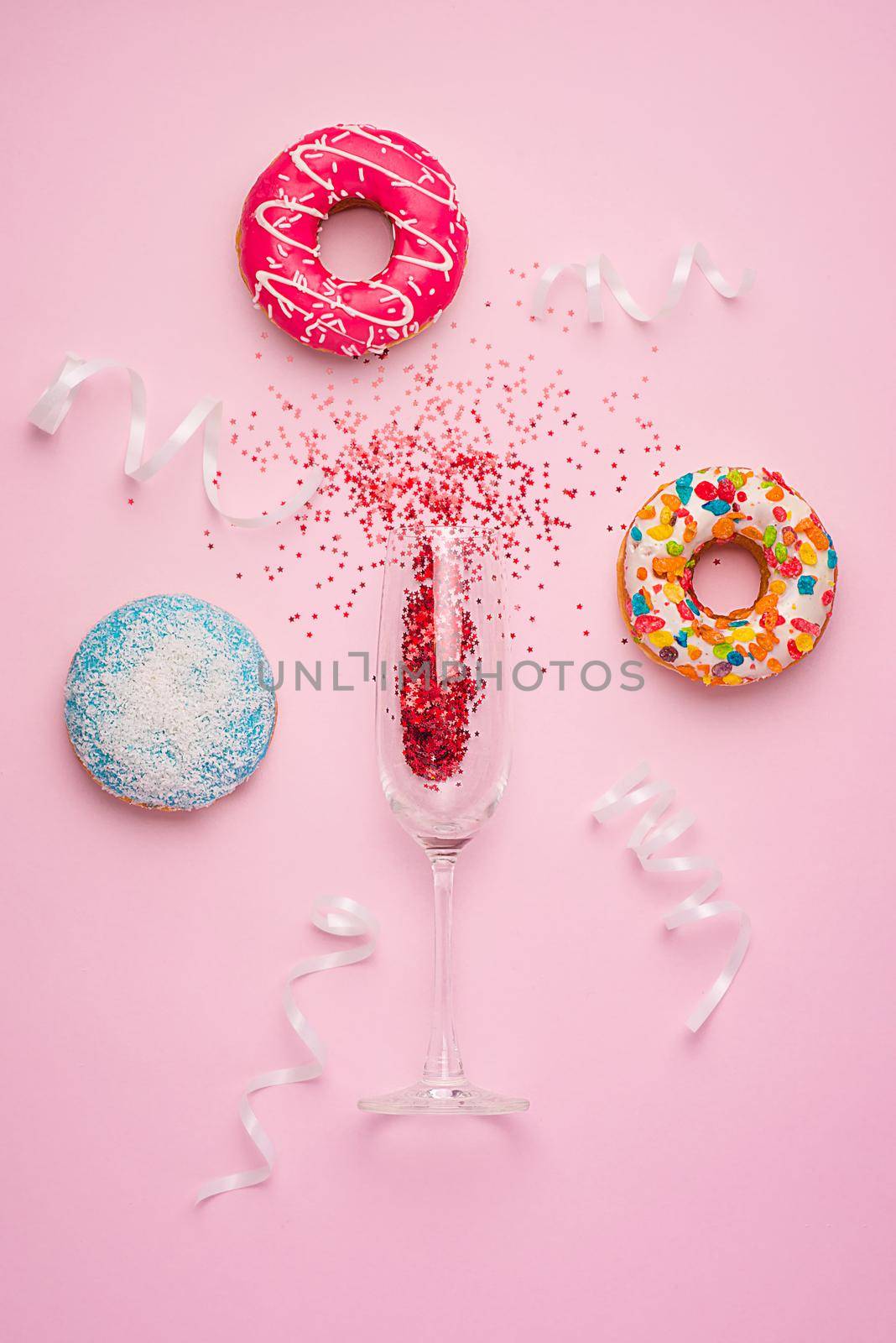 Flat lay of Celebration. Champagne glass with colorful party streamers and delicious donuts on pink background.