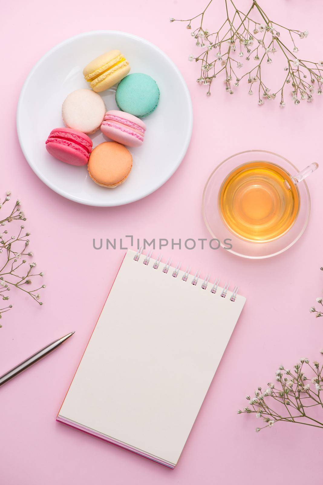 Flatlay of notebook, cake macaron, cup of tea and flower on pink table. Beautiful breakfast with macaroon.