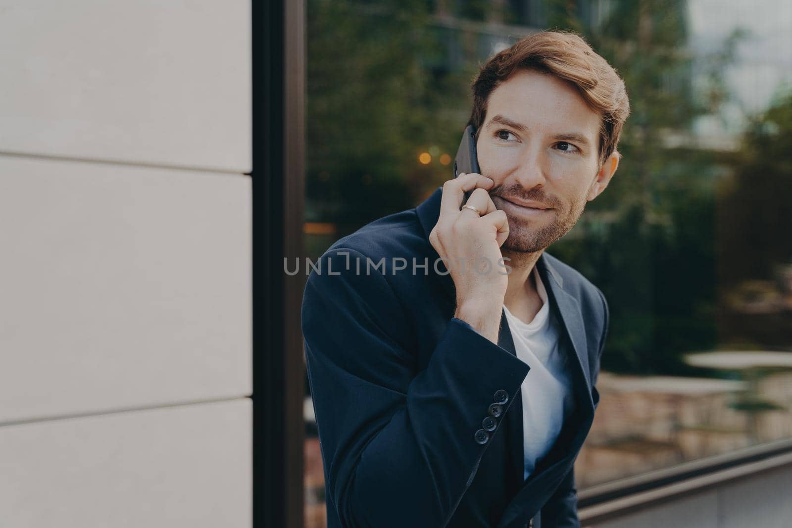 Business communication. Young handsome urban professional businessman listening to customer on cell phone, discussing something on smartphone, standing alone next to building with reflective window