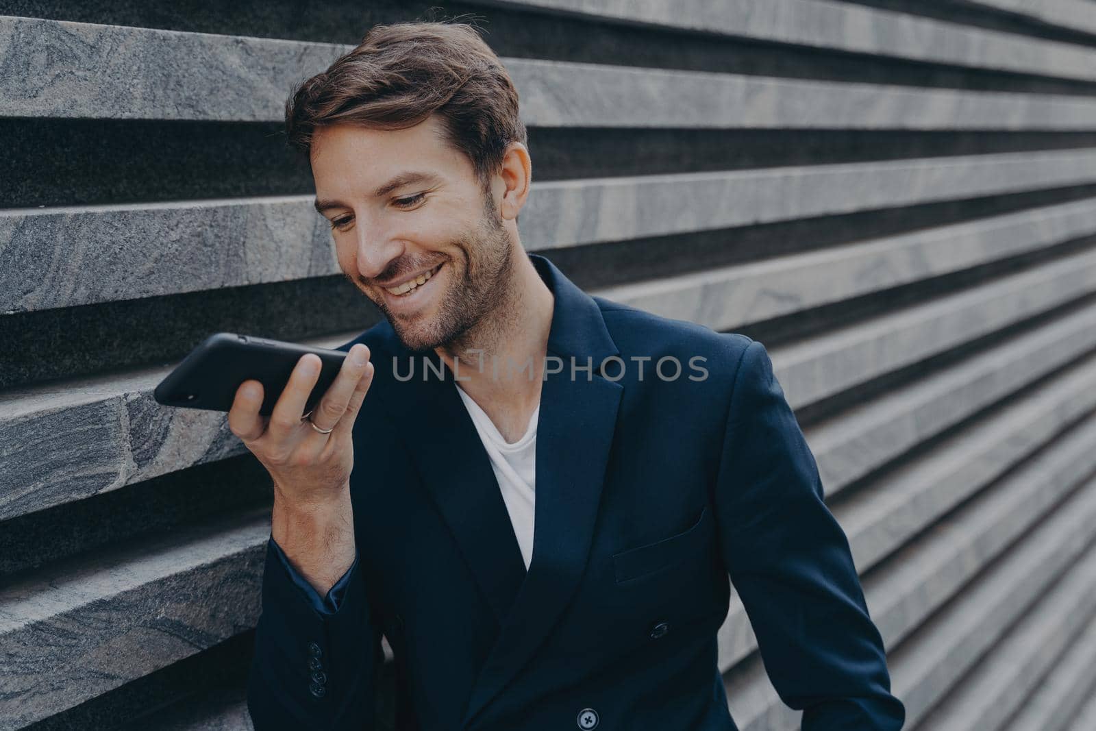 Young happy handsome man standing outside and holding phone near mouth, talking on speakerphone and smiling, recording audio message for customer or using voice recognition assistant on smartphone