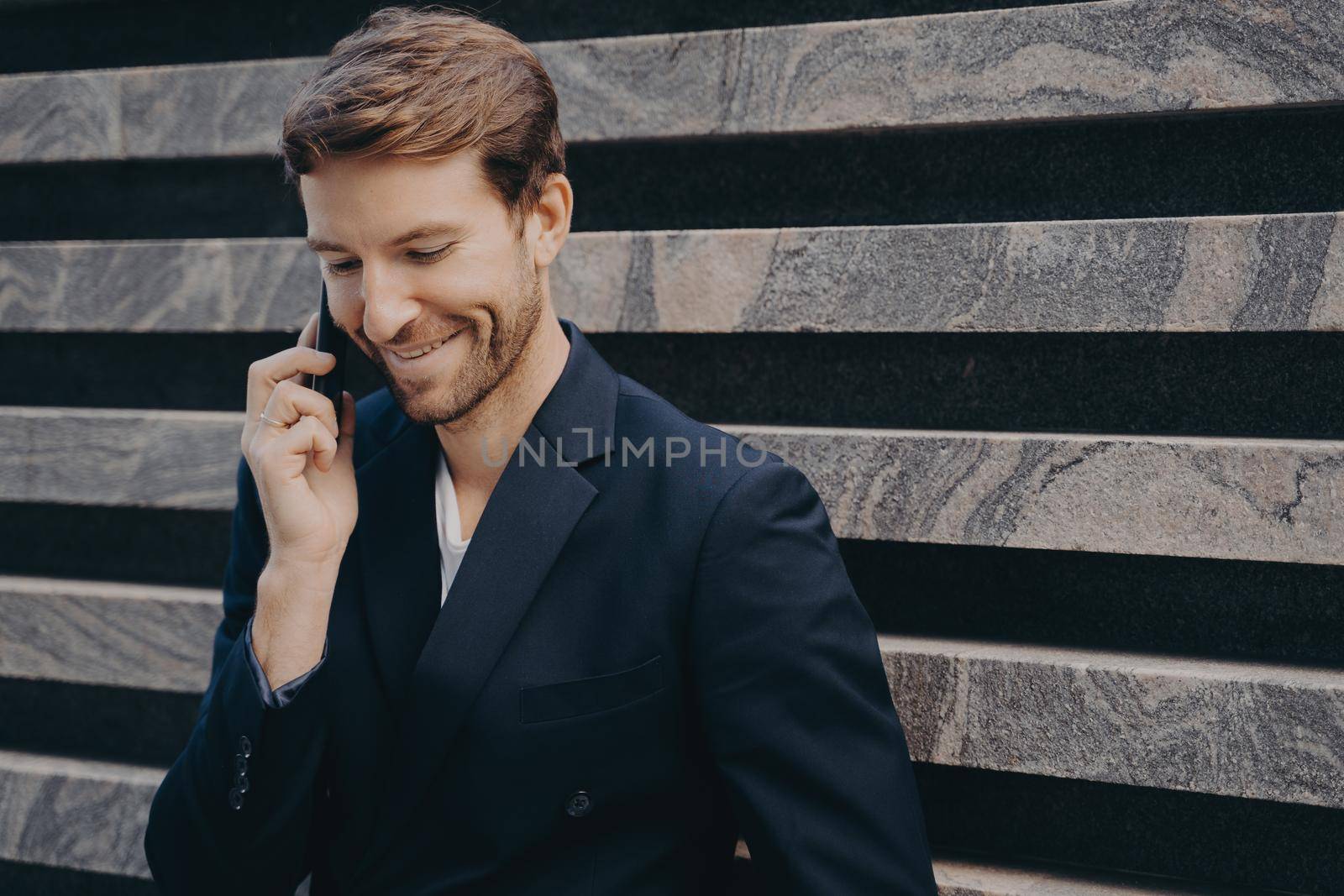 Young attractive confident businessman with neat beard in formal wear speaking on mobile leans against outer wall of city building. Smiling man having conversation on phone while having work break