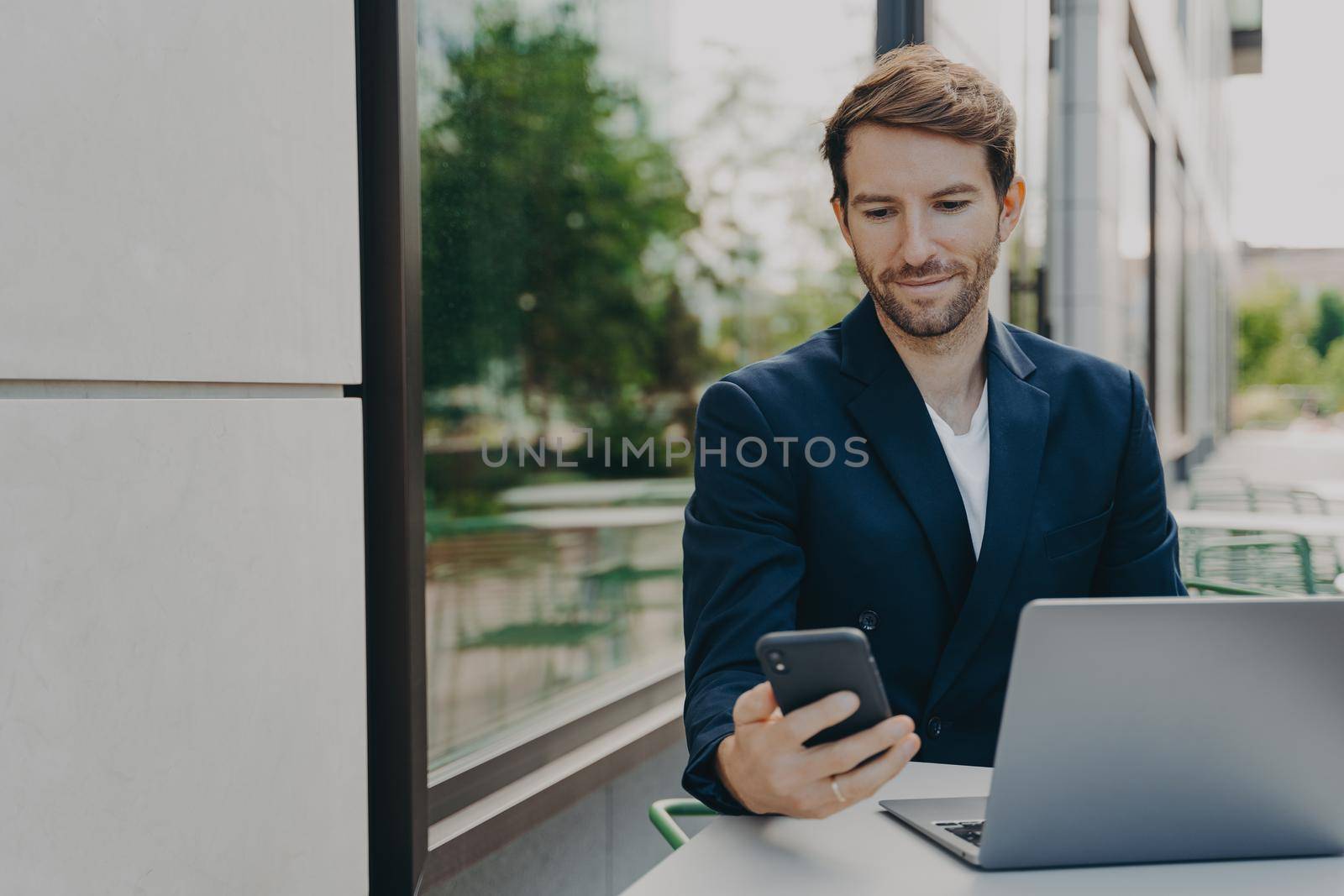 Focused young businessman working on laptop remotely, browsing phone notifications while drinking coffee outside at cafe table, male freelancer sitting at sidewalk downtown cafe and doing remote job