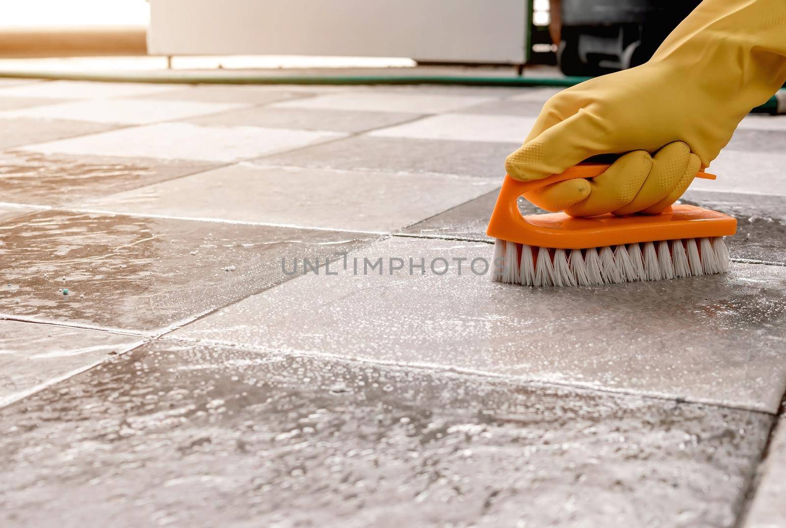Hands wearing yellow rubber gloves are using a plastic floor scrubber to scrub the tile floor with a floor cleaner.