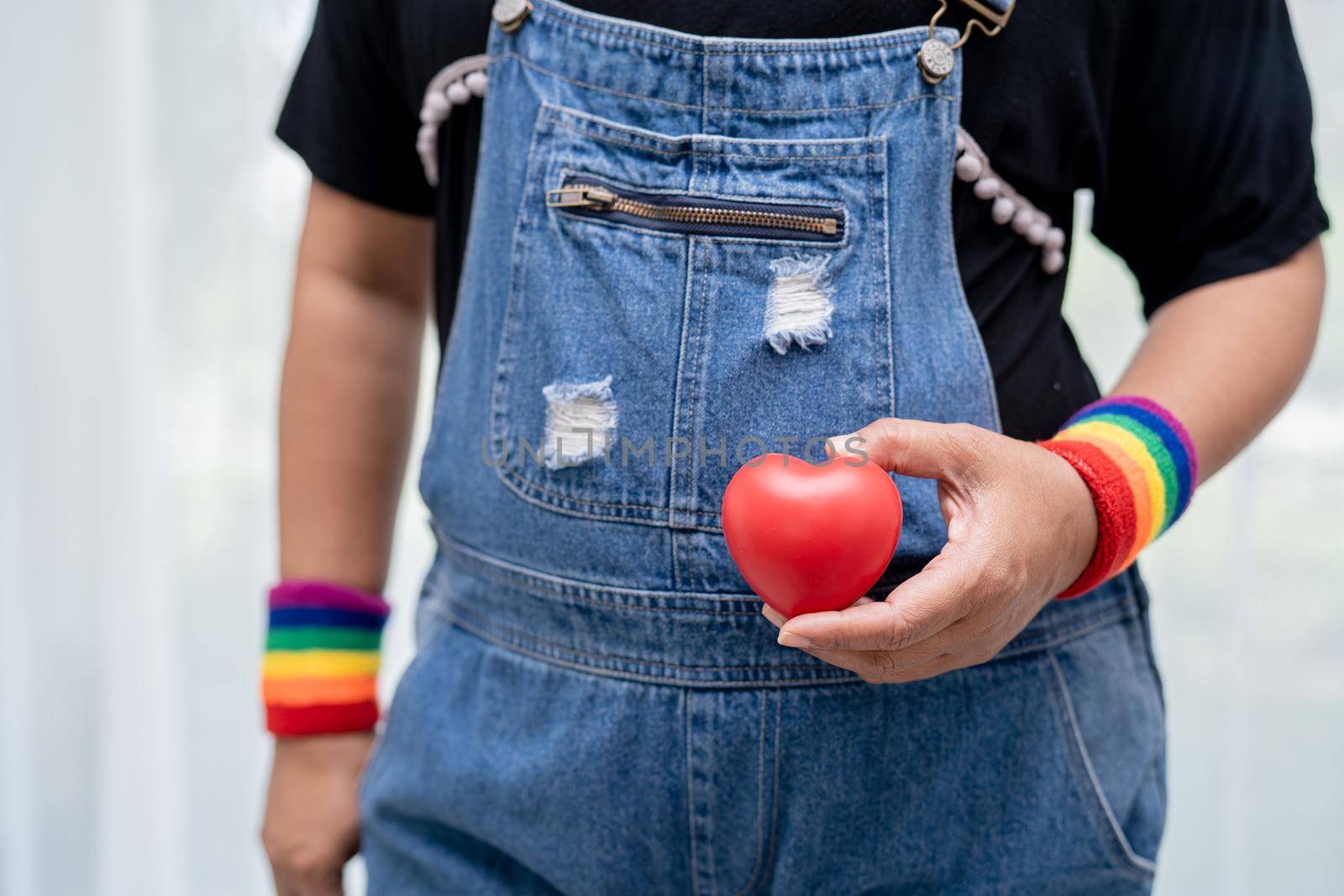 Asian lady wearing rainbow flag wristbands and hold red heart, symbol of LGBT pride month celebrate annual in June social of gay, lesbian, bisexual, transgender, human rights.