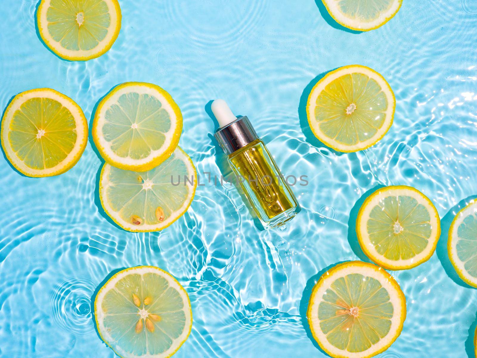 Luxury cosmetic oil or serum in glass bottle with pipette in clean transparent water with lemon slices over blue background. Water splashing on water surface in sunlight. Vertical flatlay copy space