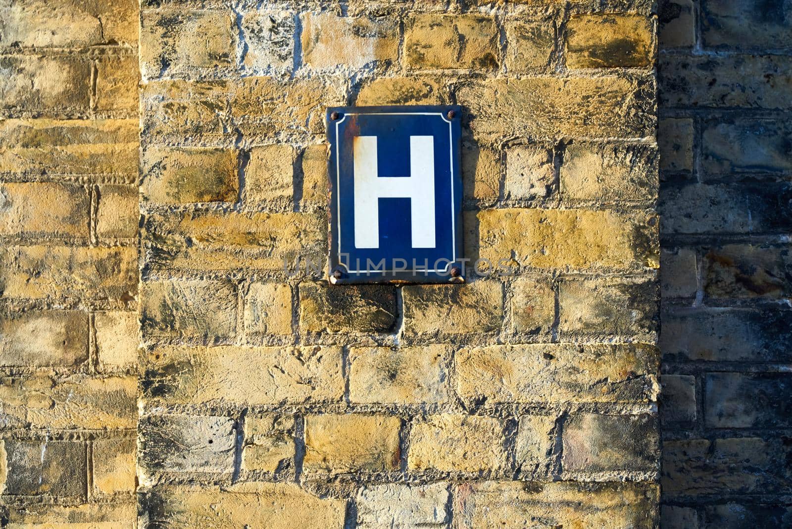 A white letter H on a blue metal plate mounted on a yellow brick wall