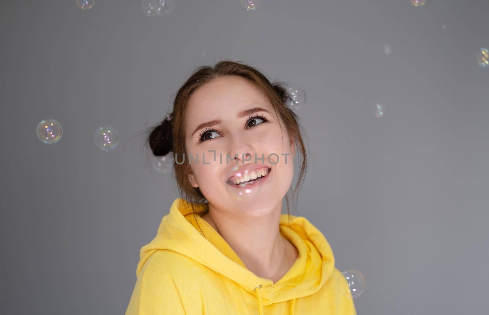 happy brunette woman in yellow hoodie among soap bubbles in bright grey room. happy people. millennial generation. fashionable teenager. trendy colours