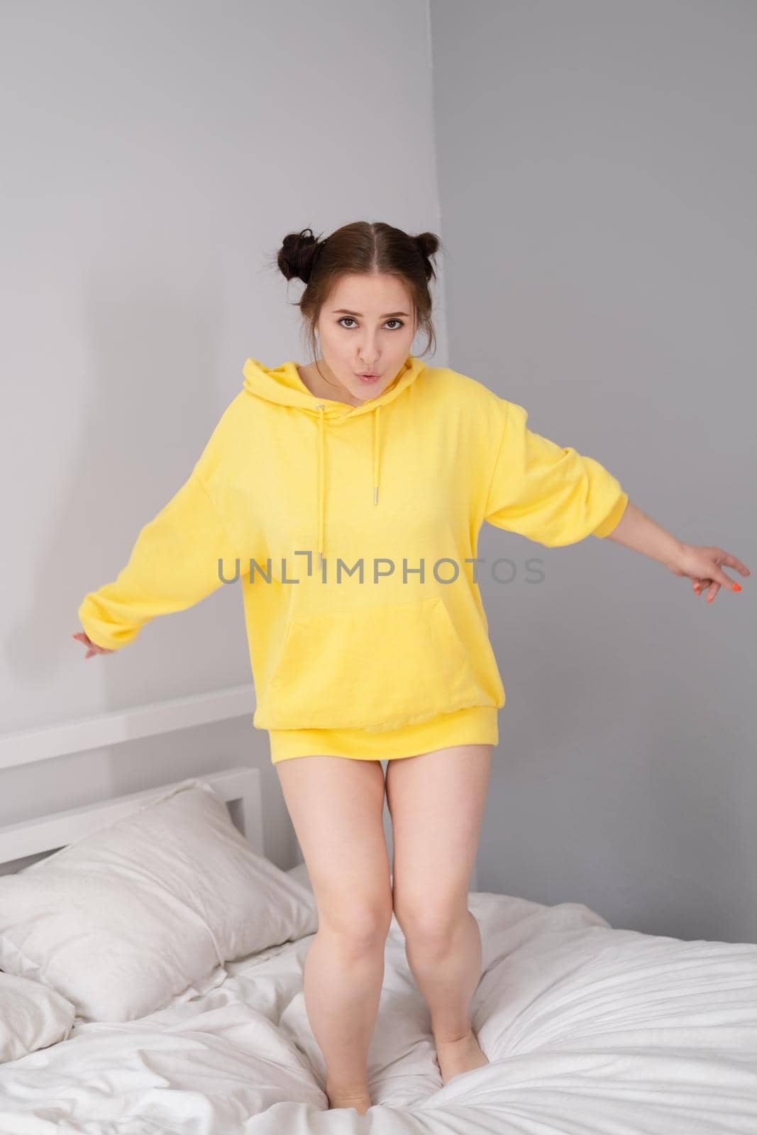 cheerful positive brunette woman in yellow hoodie in white bedroom on bed with white linens. happy people. millennial generation. fashionable teenager.