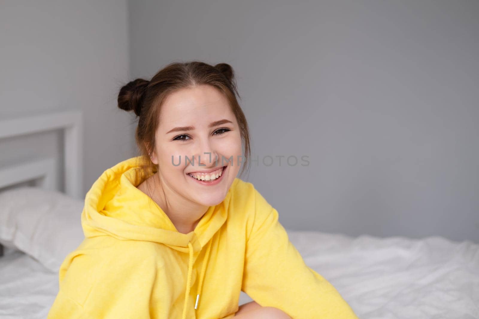 happy cheerful brunette girl in yellow hoodie in white bedroom sitting on bed with white bed linens