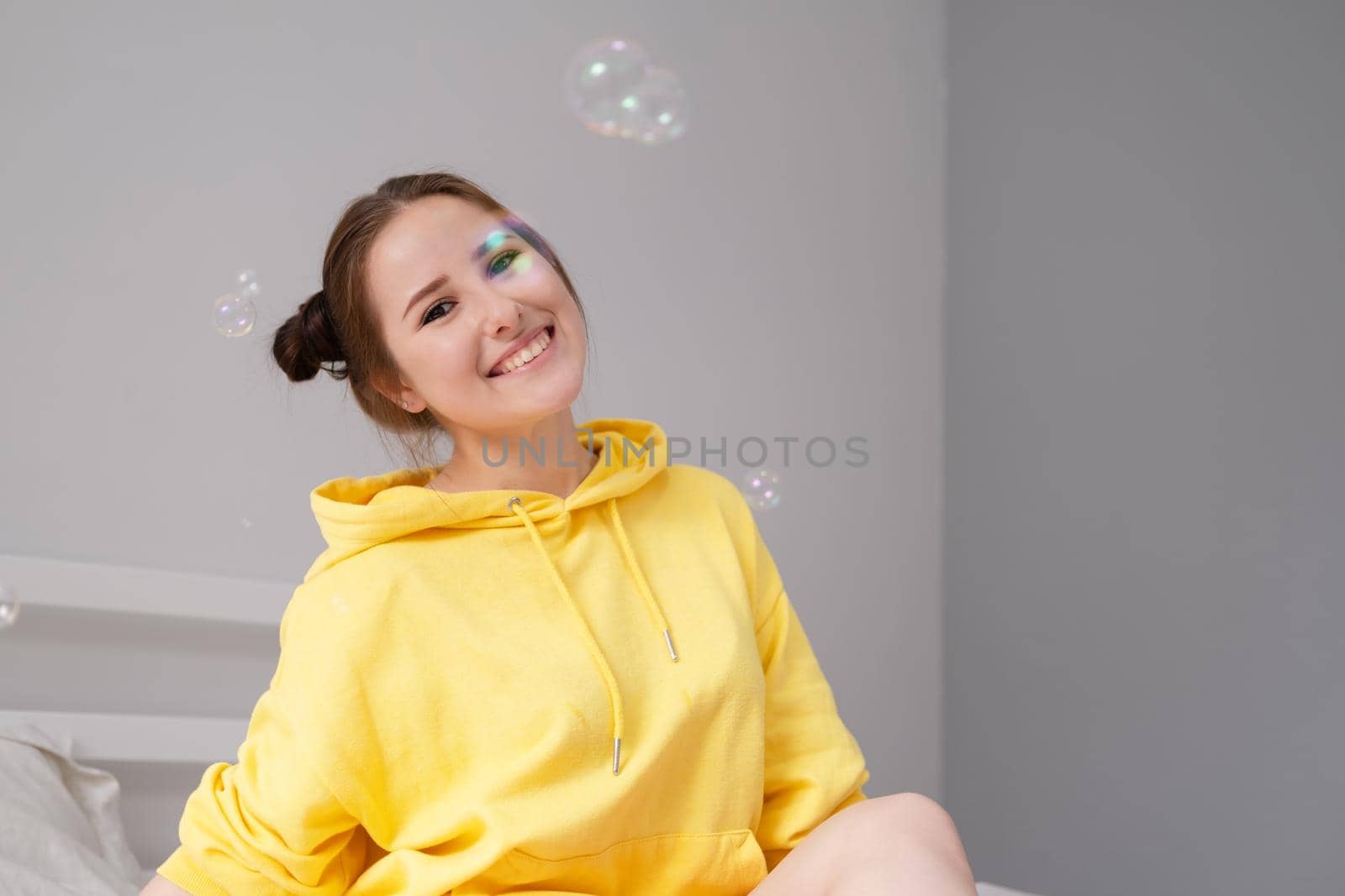 happy brunette woman in yellow hoodie among soap bubbles in bright grey room. happy people. millennial generation. fashionable teenager. trendy colours