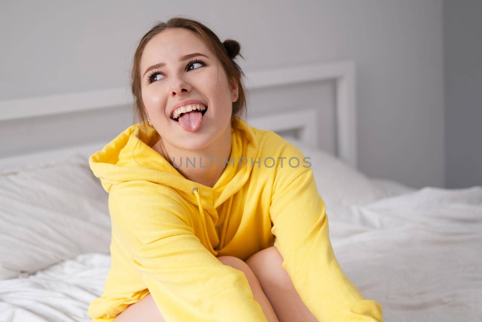 cheerful positive brunette woman in yellow hoodie in white bedroom on bed with white linens. happy people. millennial generation. fashionable teenager.