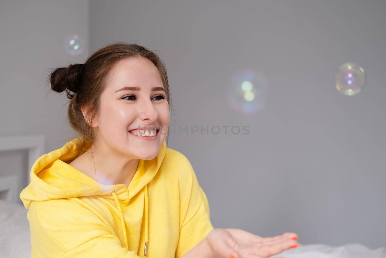 happy brunette woman in yellow hoodie among soap bubbles in bright grey room. happy people. millennial generation. fashionable teenager. trendy colours