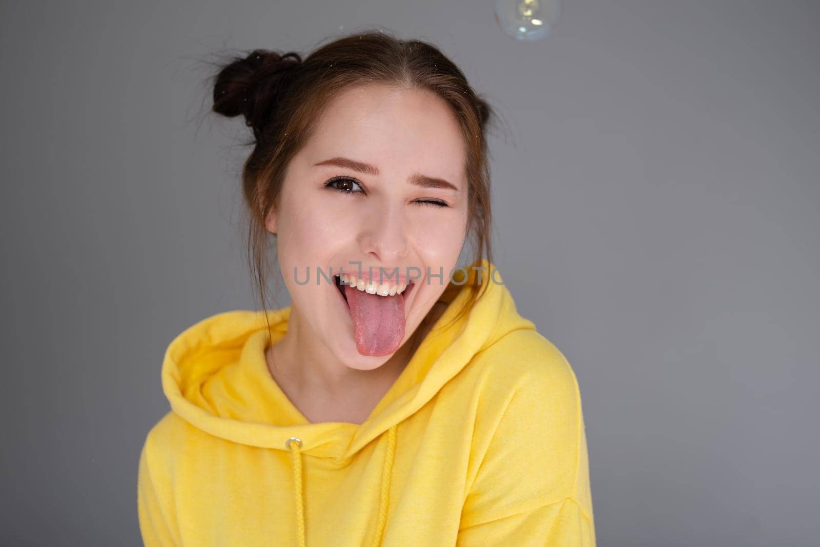 happy brunette woman in yellow hoodie among soap bubbles in bright grey room. happy people. millennial generation. fashionable teenager. trendy colours