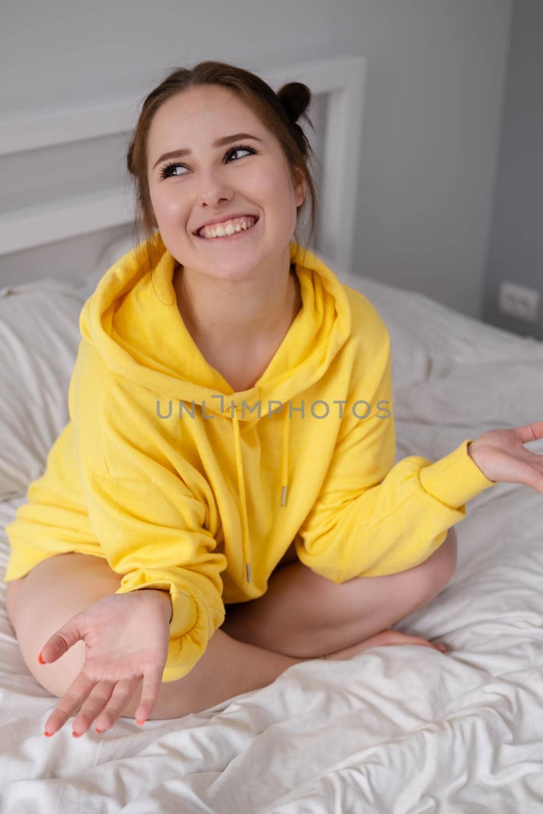 happy cheerful brunette girl in yellow hoodie in white bedroom sitting on bed with white bed linens. by oliavesna