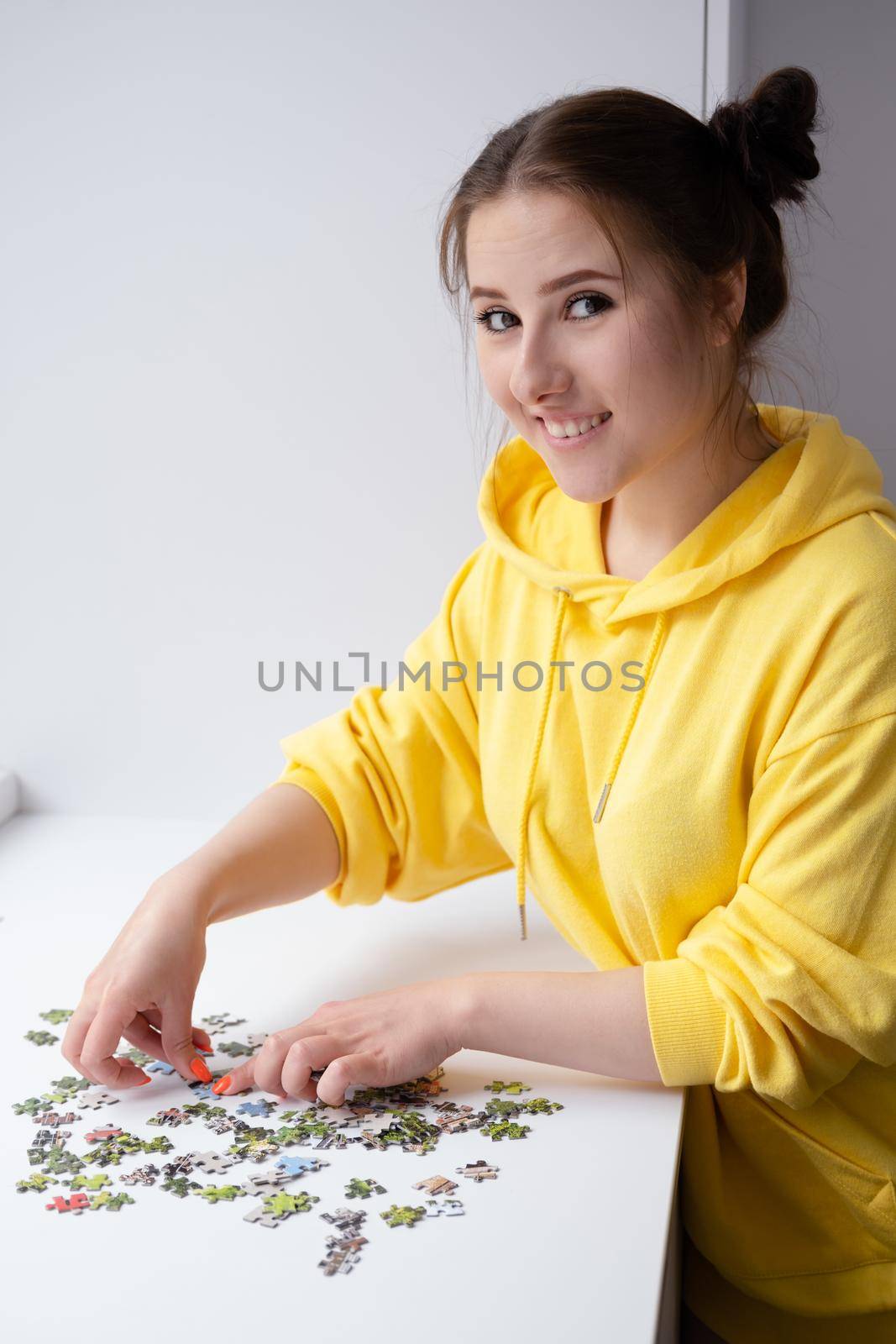 pretty brunette girl in yellow hoodie arranging pieces of a Jigsaw Puzzle Game in bright white room. playing board game home. by oliavesna