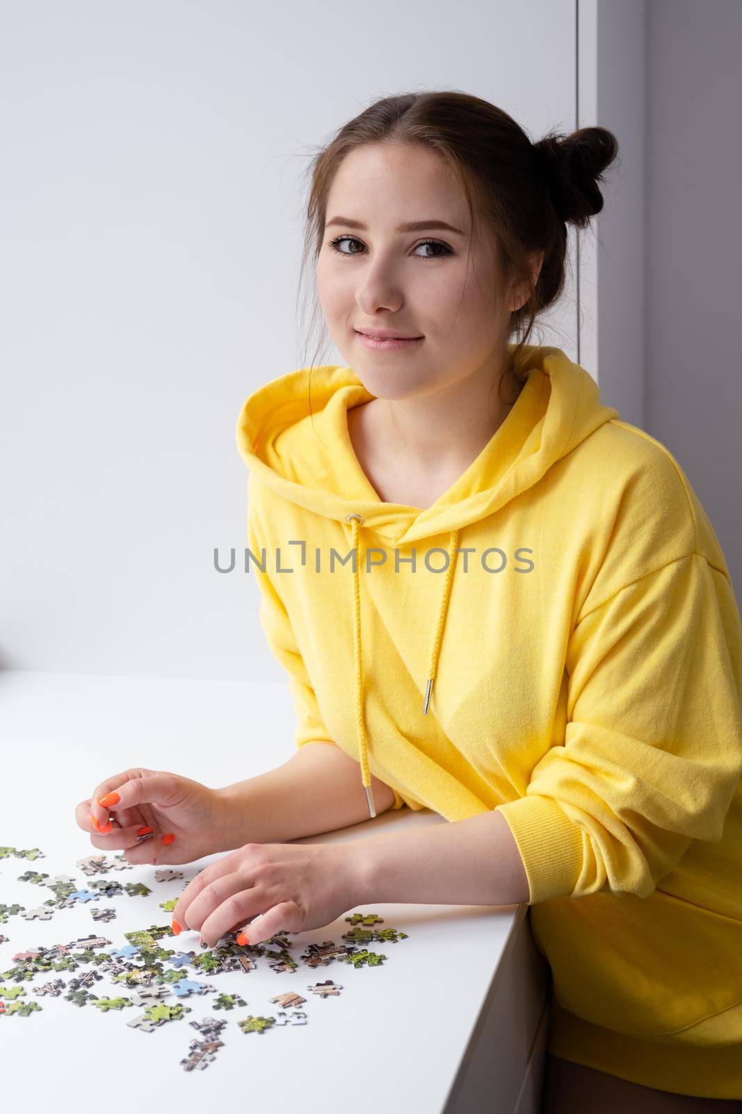 pretty brunette girl in yellow hoodie arranging pieces of a Jigsaw Puzzle Game in bright white room. playing board game home