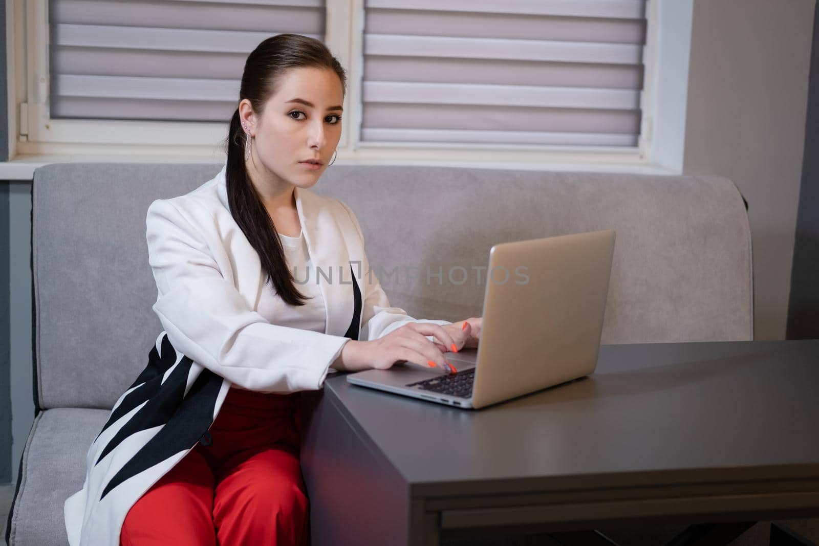 woman sitting by the table on couch in grey room - office and use laptop. big boss. important, confident women. femininity. millennials.