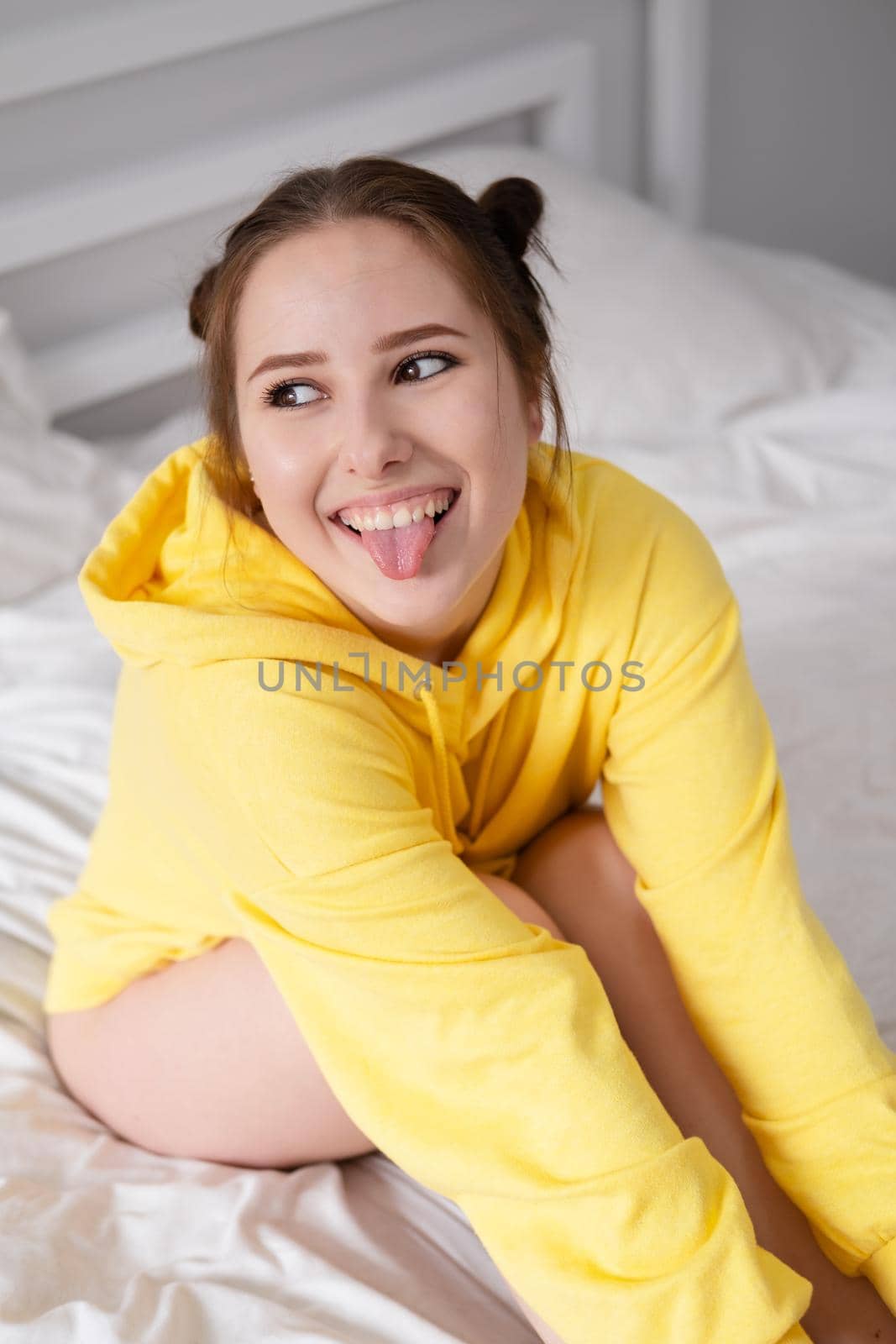 cheerful positive brunette woman in yellow hoodie in white bedroom on bed with white linens. happy people. millennial generation. fashionable teenager by oliavesna