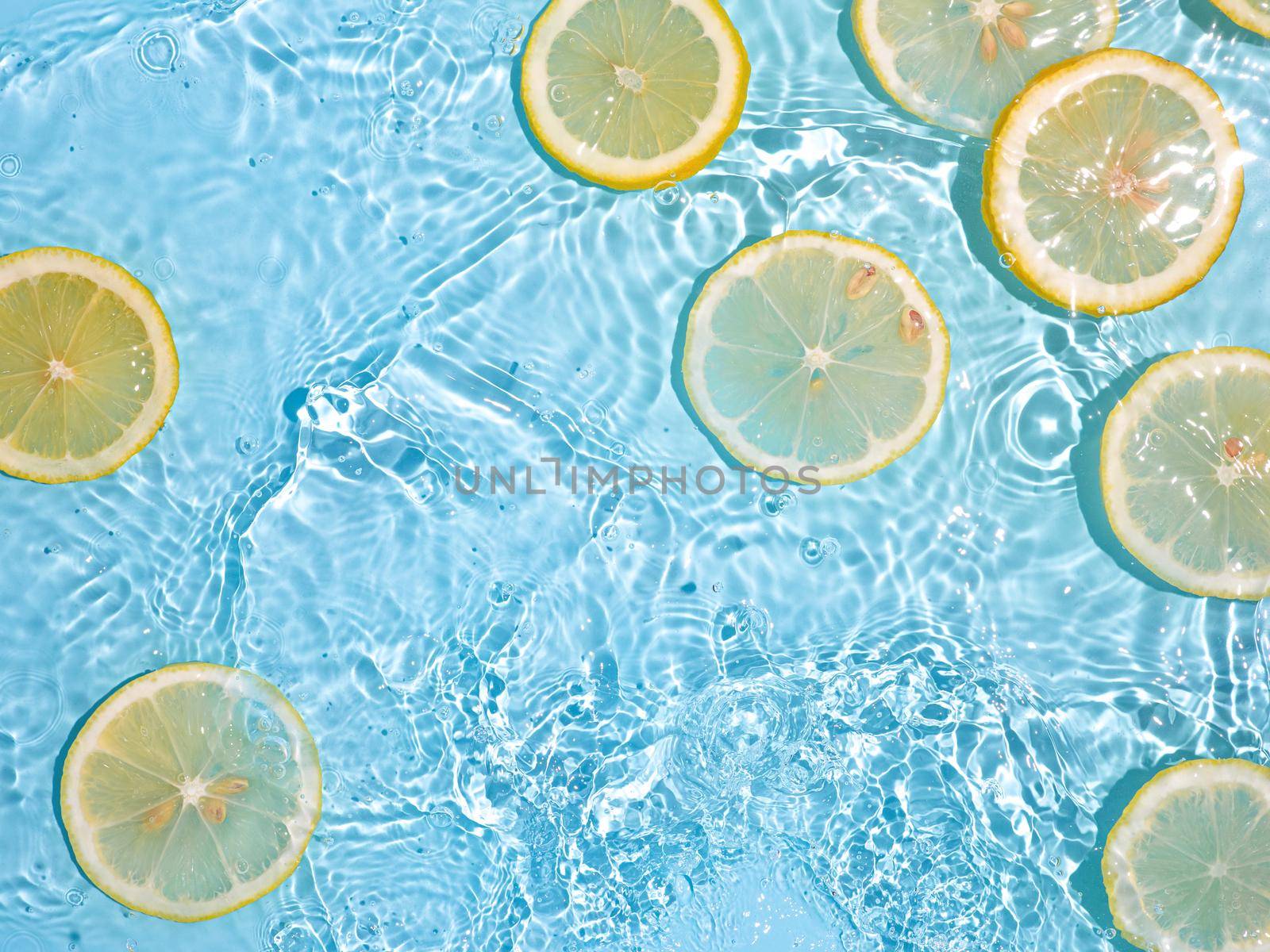 Lemon slices in clean transparent water over blue background with copy space. Water splashing on blue water surface in sunlight. Top view or flatlay. Summer, vacation, healthy eating concept