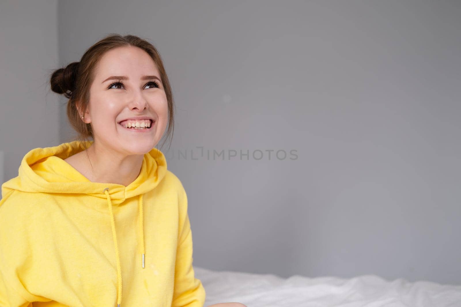 cheerful positive brunette woman in yellow hoodie in white bedroom on bed with white linens. happy people. millennial generation. fashionable teenager.