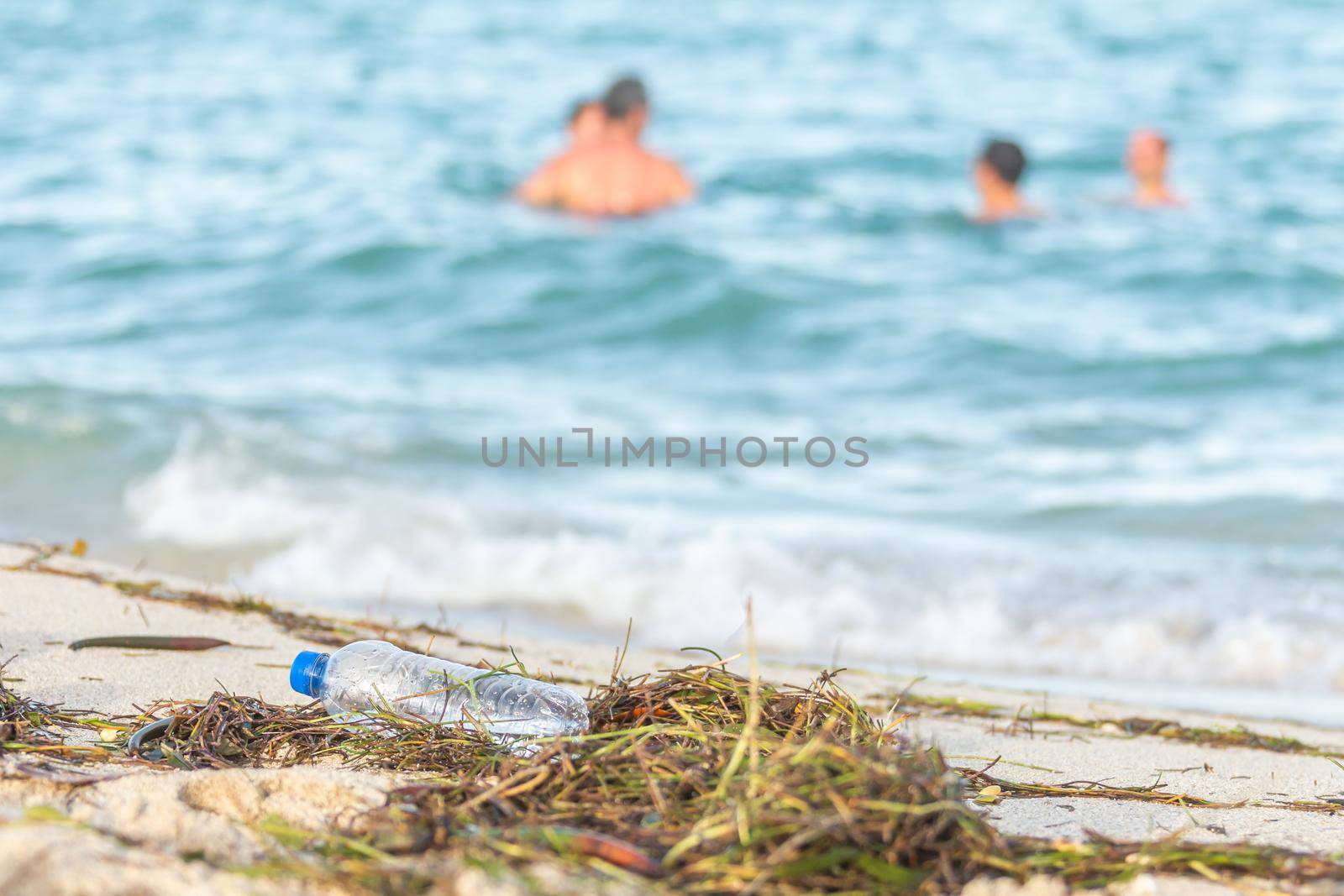 close up image of empty plastic water bottle on dirty beach filled with seaweed on dirty sand beach with people in sea on background