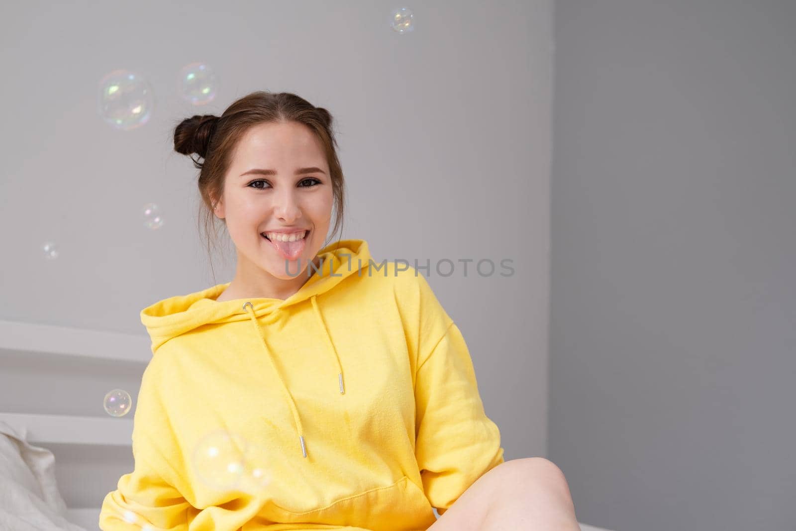 cheerful positive brunette woman in yellow hoodie in white bedroom on bed with white linens. happy people. millennial generation. fashionable teenager.