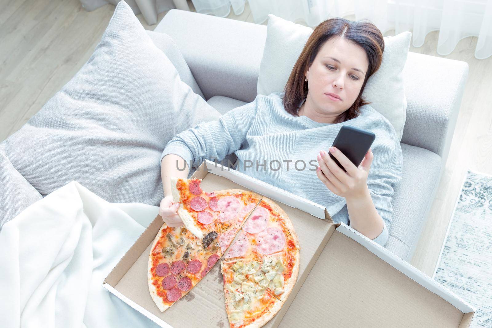 Sad woman eating pizza and holding phone laying on a couch at home