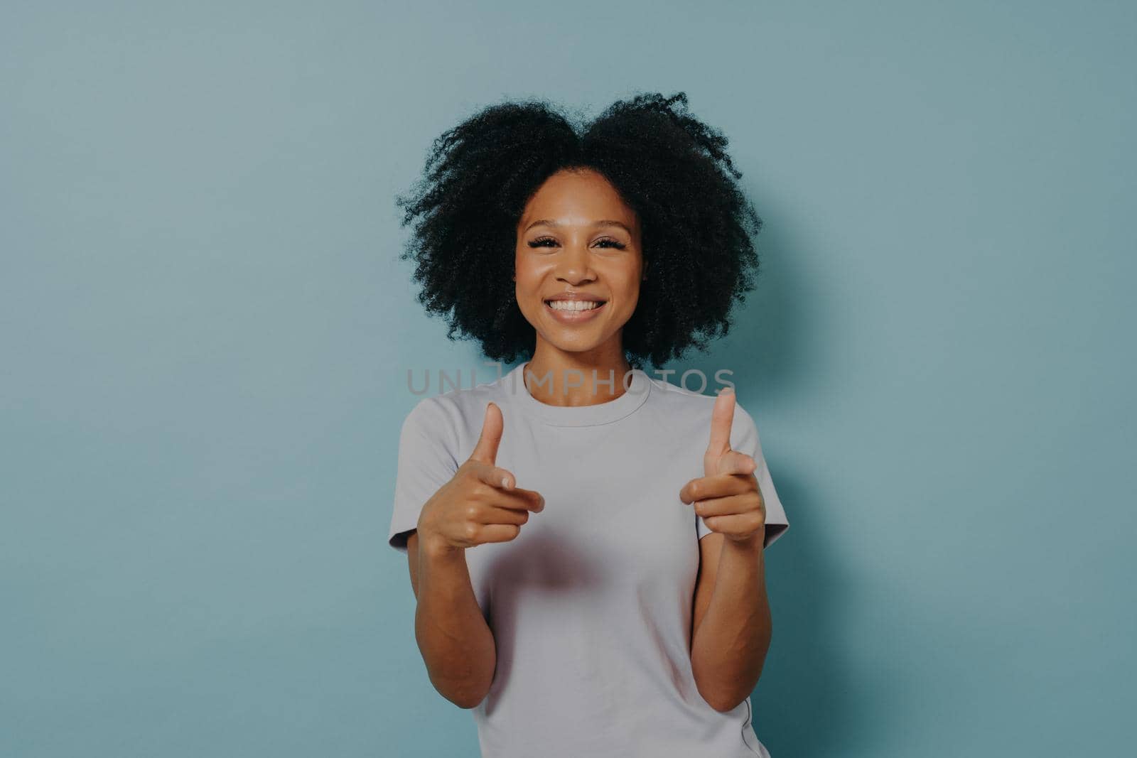Positive friendly dark skinned lady making finger gun gesture and smiling at camera, happy young african female in white tshirt being happy to meet best friend, isolated over blue background