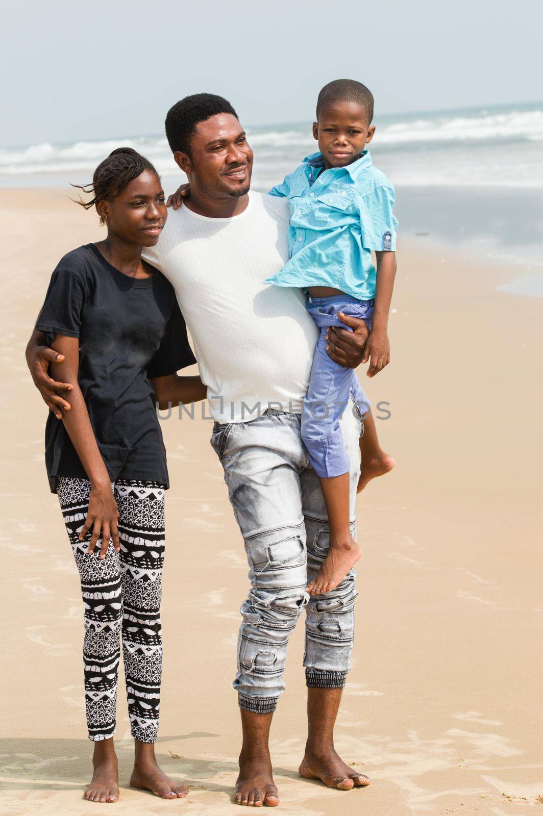 Young man at the beach with his children. He lifts the youngest, and the right hand holding the biggest by his shoulder all standing at the beach.