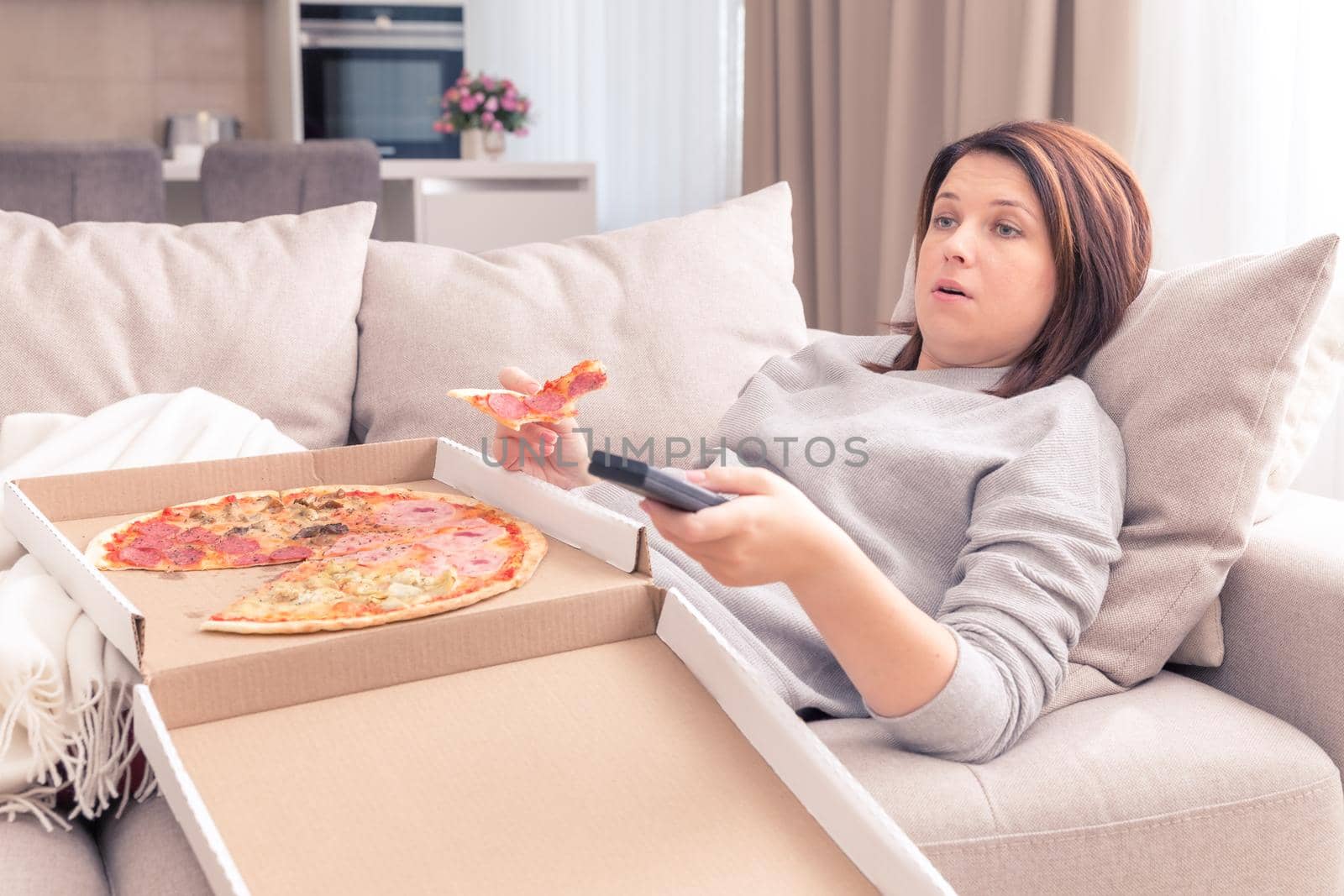 Beautiful woman eating pizza and watching TV holding remote control at home