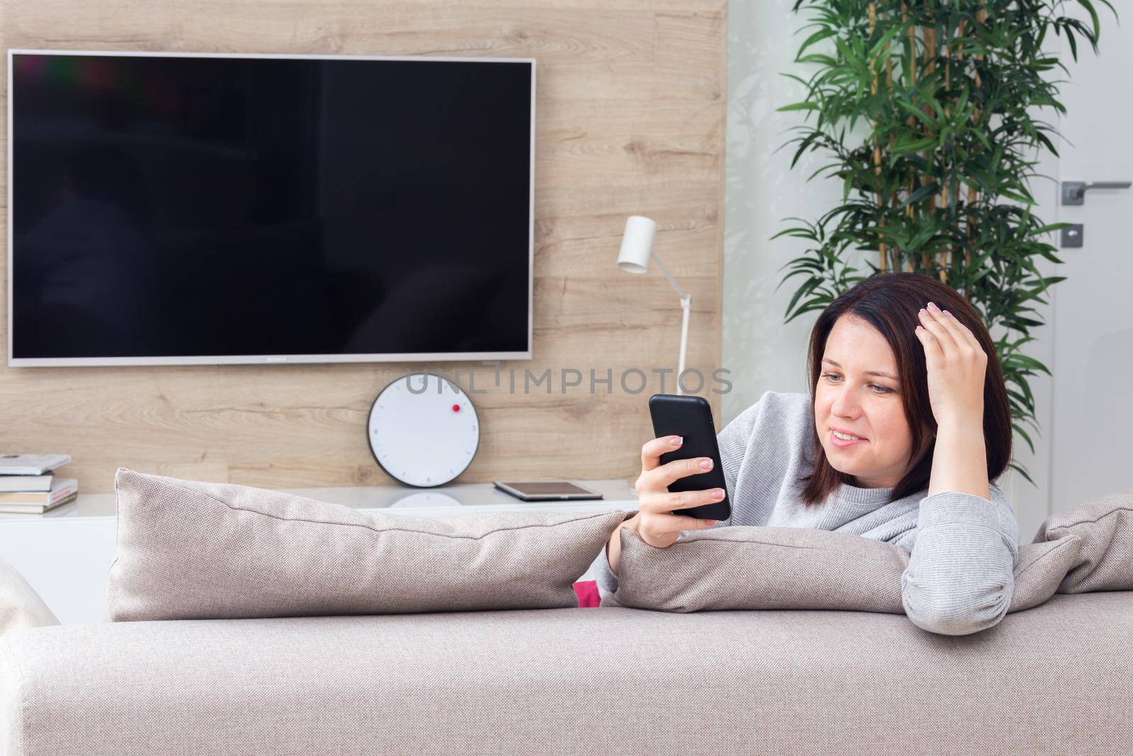 Young woman looking at her smart phone and smiling while sitting on the couch at home