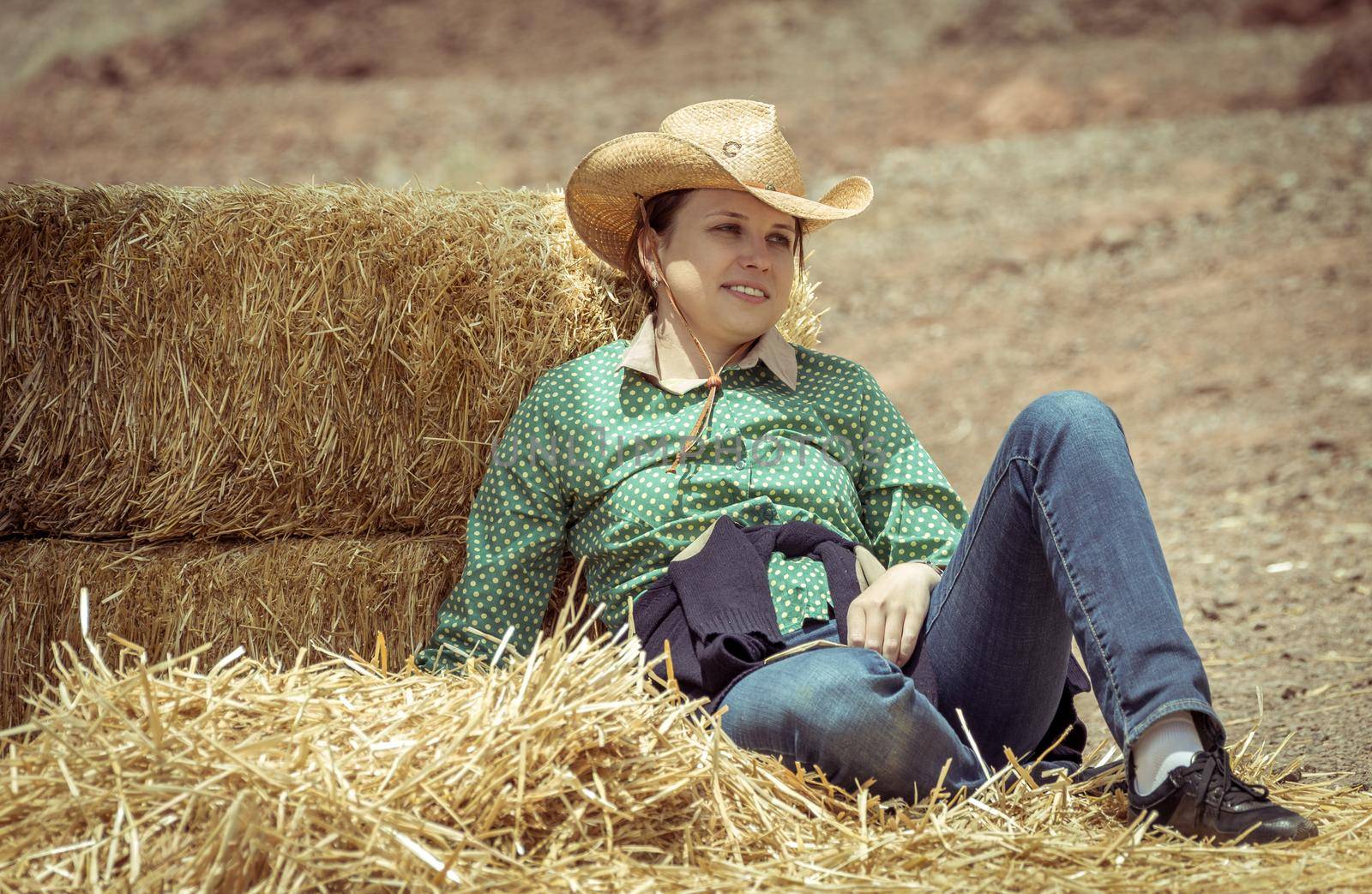 Cowgirl woman smiling happy sitting next to haystack wearing cowboy hat. Beautiful young Caucasian girl in countryside