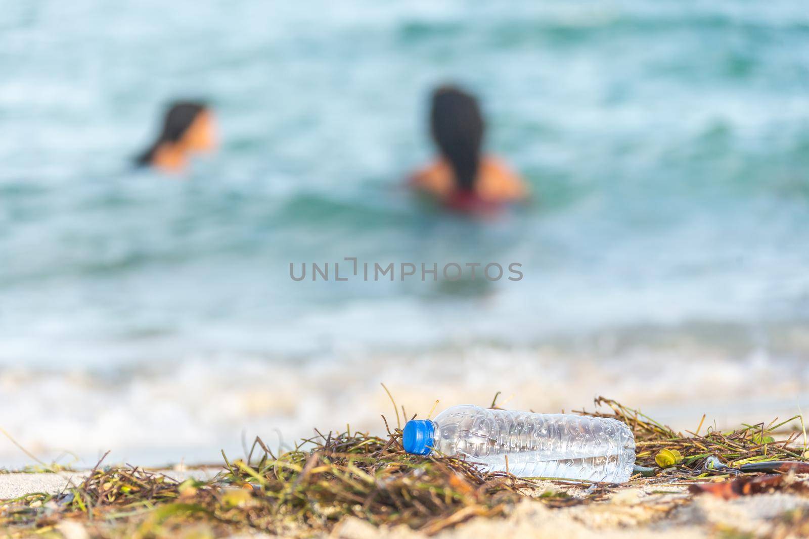 close up image of empty plastic water bottle on dirty beach filled with seaweed on dirty sand beach with people in sea on background
