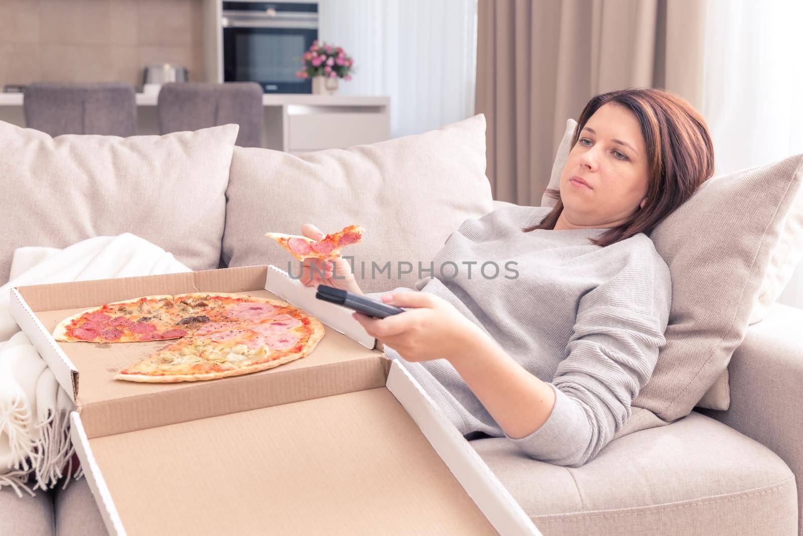 Sad woman eating pizza and holding phone laying on a couch at home