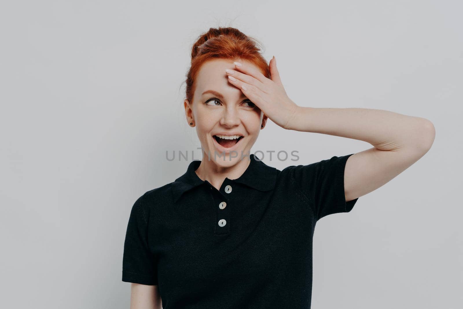 Happy young redhead woman with opened mouth touching her forehead with hand, smiling and looking aside while standing against grey wall in studio, trying to remember something, dressed in black tshirt