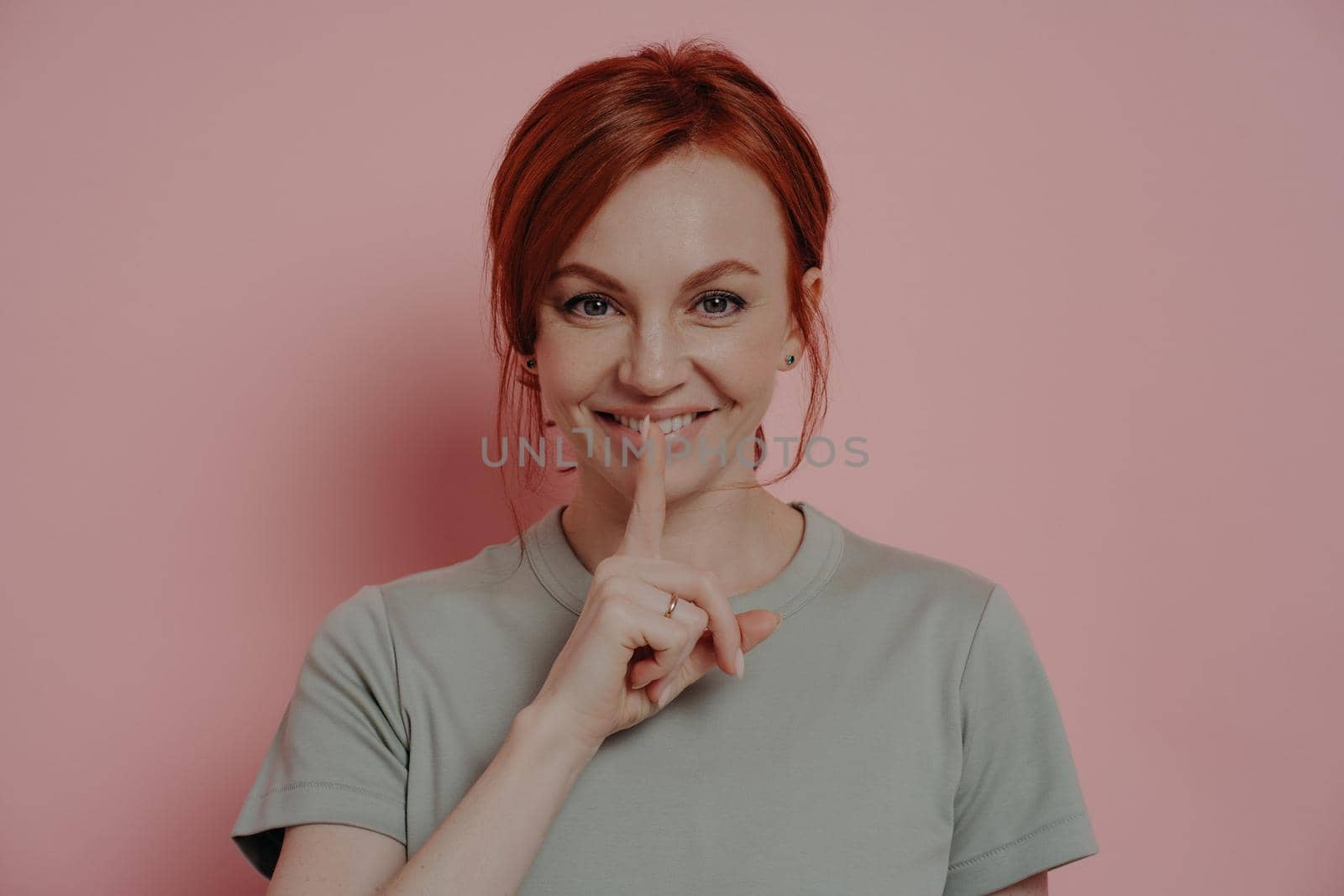 Young beautiful smiling red-haired woman telling secret information, posing isolated over pink wall by vkstock