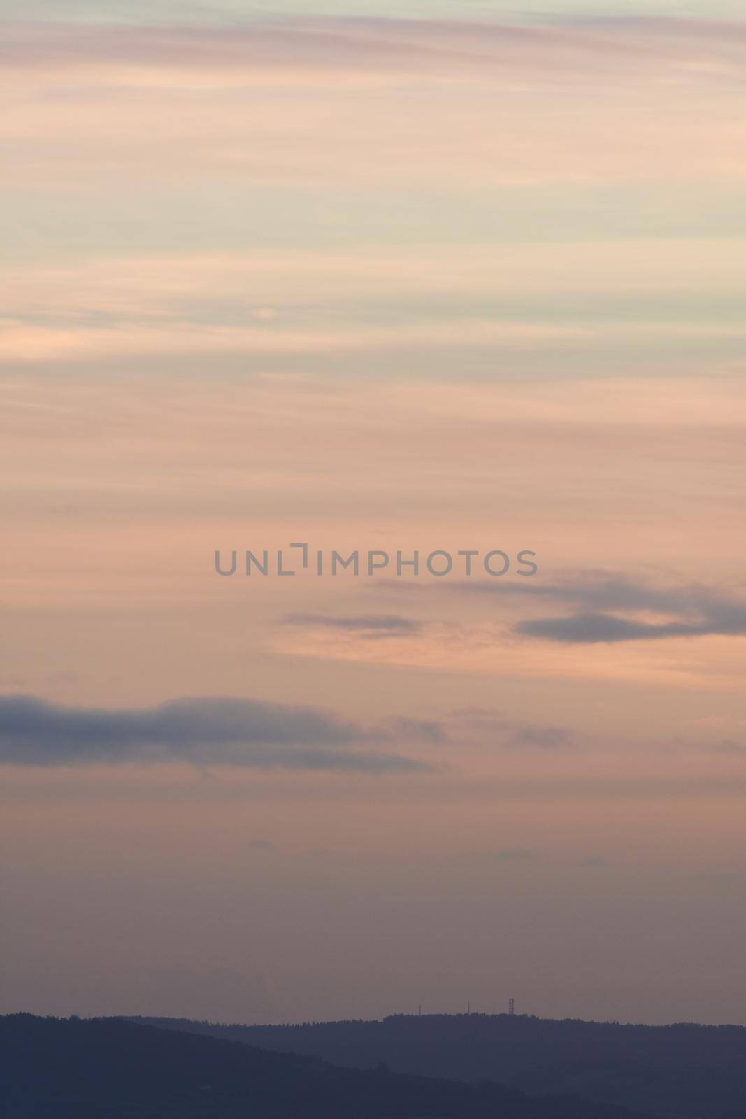 Vertical hazy sunset. Horizon under warm and faded pastel colours. Gently clouded sky above the forested horizon with protruding telecom towers.