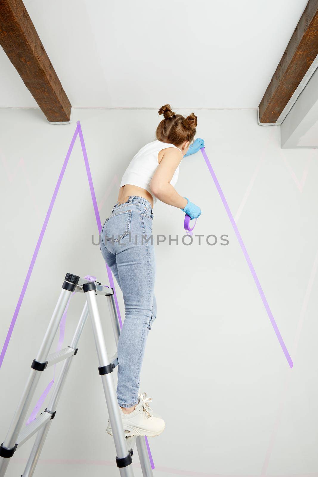 Young woman preparing wall for painting standing on ladder applying masking tape on wall. Diy project by Mariakray