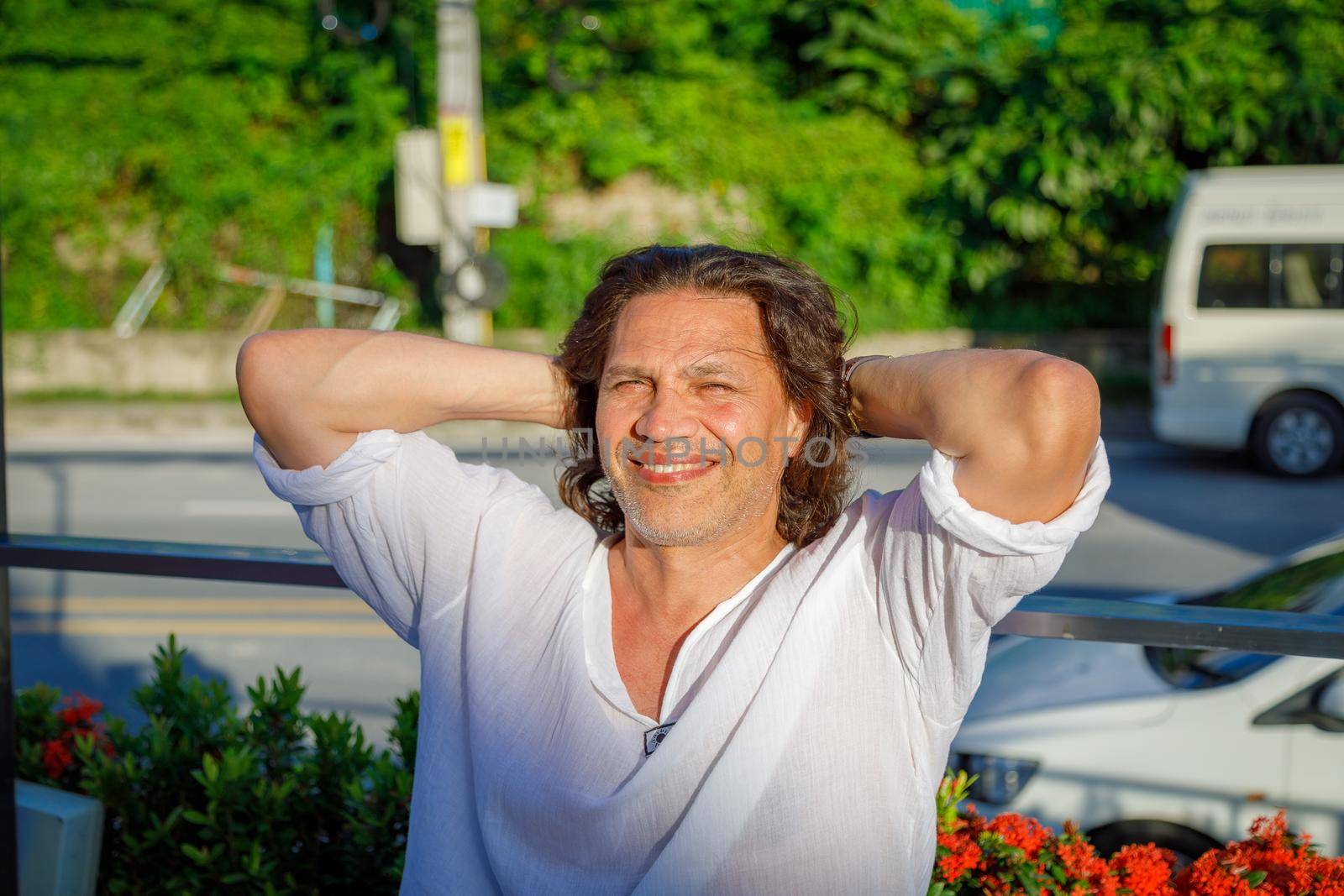 Happy adult man with long hair smiling, throwing his hands behind his head. The man in the white shirt is happy with the vacation.