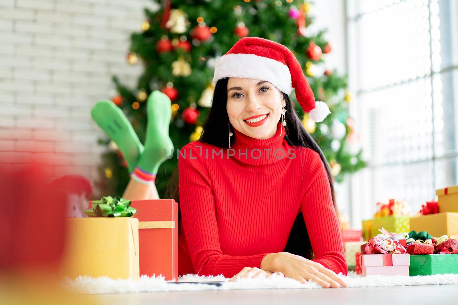 Pretty woman with Christmas costume lie on floor and look at camera with smiling stay at home with the present and Christmas tree as background.